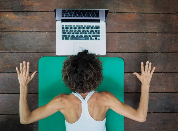Evde yoga yapan bir kadın dizüstü bilgisayarın önünde oturuyor.