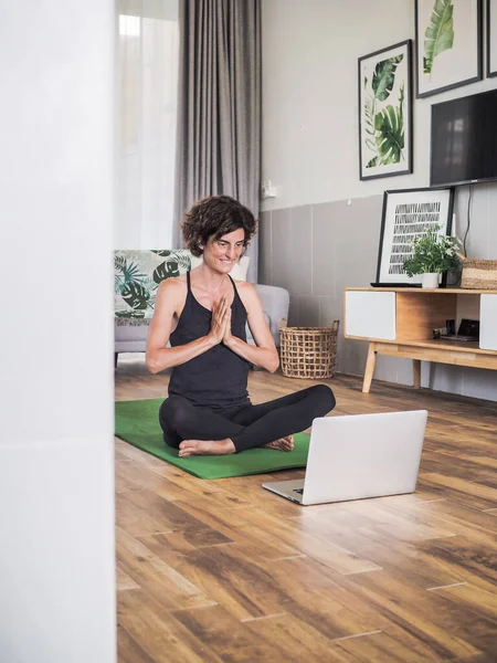 Evde yoga yapan bir kadın dizüstü bilgisayarın önünde oturuyor.