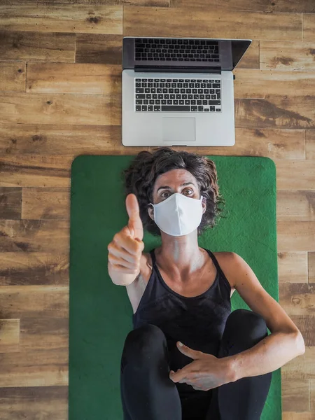 emotional woman in mask lying on yoga mat on floor with laptop behind her head