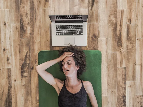 sleepy woman lying on yoga mat on floor with laptop behind her head