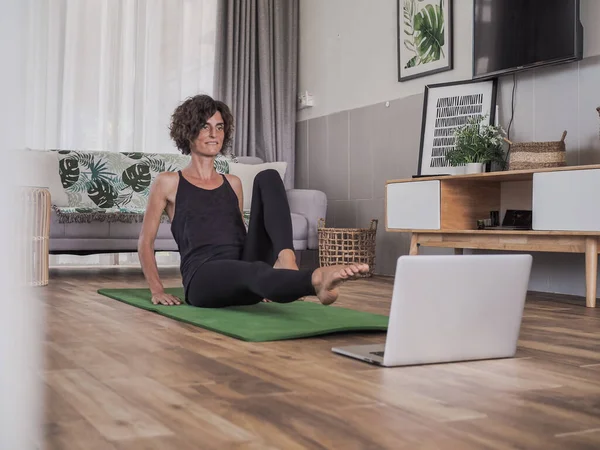 Evde yoga yapan bir kadın dizüstü bilgisayarın önünde oturuyor.