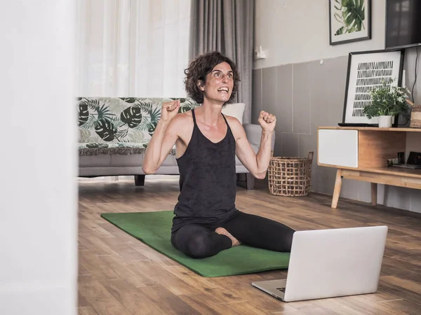 Evde yoga yapan bir kadın dizüstü bilgisayarın önünde oturuyor.