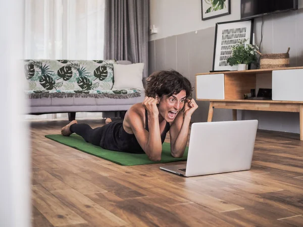 Evde dizüstü bilgisayar kullanan mutlu bir kadın yoga minderinde yatıyor.