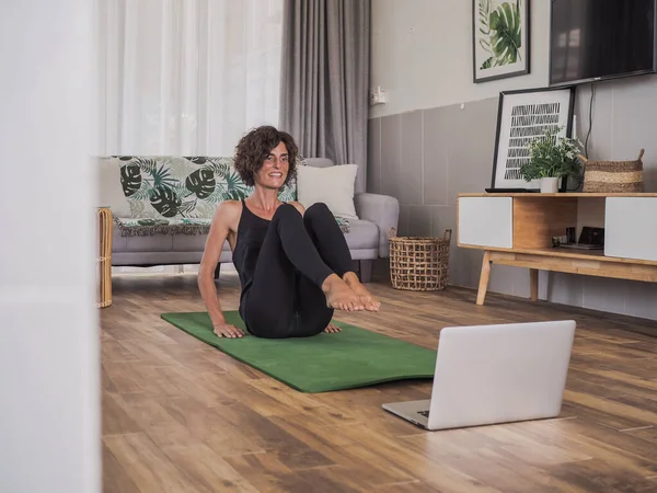 Evde yoga yapan bir kadın dizüstü bilgisayarın önünde oturuyor.