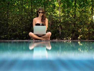 bikinili genç bir kadın havuzun kenarında dizüstü bilgisayarla oturuyor.