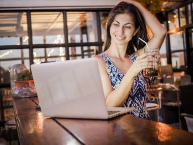 Kafede dizüstü bilgisayar kullanan, kokteyl içen mutlu genç bir kadın.
