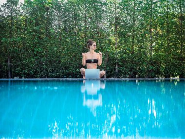 bikinili genç bir kadın havuzun kenarında dizüstü bilgisayarla oturuyor.