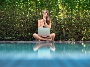 bikinili genç bir kadın havuzun kenarında dizüstü bilgisayarla oturuyor.