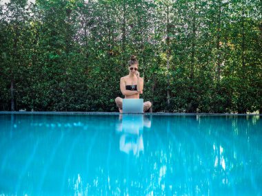 bikinili genç bir kadın havuzun kenarında dizüstü bilgisayarla oturuyor.