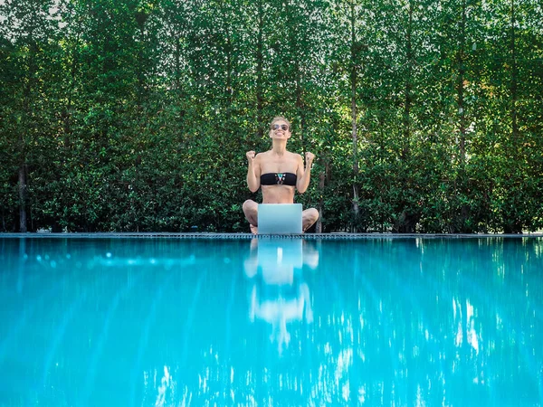 young woman in bikini sitting on the edge of the pool with laptop