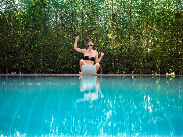 young woman in bikini sitting on the edge of the pool with laptop