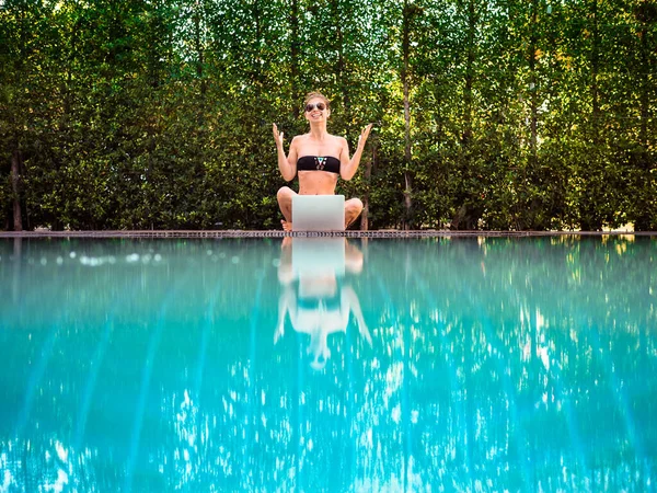 young woman in bikini sitting on the edge of the pool with laptop