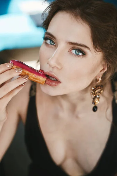 Portrait of beautiful and fashionable brunette model girl with blue eyes and with gentle makeup, with big earrings and gold manicure eating eclair and posing at interior