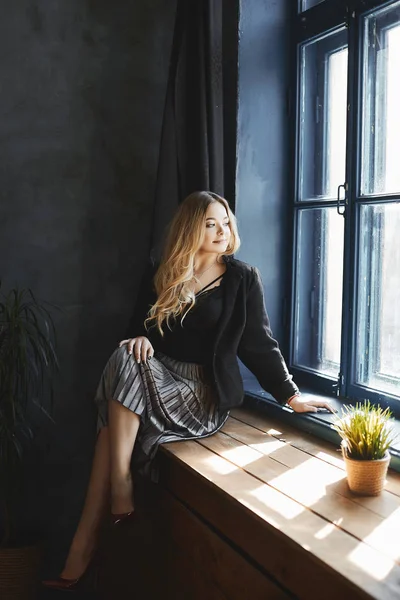 Красива і модна блондинка модель дівчина з великими грудьми в срібній спідниці і в чорній атласною блузці сидить на дерев'яному підвіконні і дивиться у вікно — стокове фото