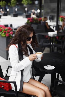 Seksi ve şık esmer modeli kız profesyonel makyaj ve şık güneş gözlüğü, beyaz şort ve beyaz bir ceket ile kahve ve kırmızı debriyaj çanta kafede masada oturur