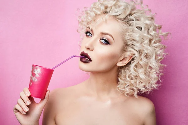 Moderiktiga och vackra blond modell flicka med blå ögon och med professionell ljusa makeup, drycker av halm från en rosa glas och poserar på studio på rosa bakgrund — Stockfoto
