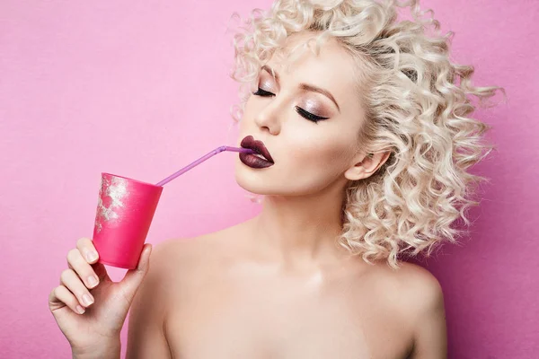 Menina modelo loira elegante e bonita com olhos fechados e com maquiagem profissional brilhante, ela bebe por uma palha de um copo rosa e posando em estúdio em fundo rosa. Instrução — Fotografia de Stock