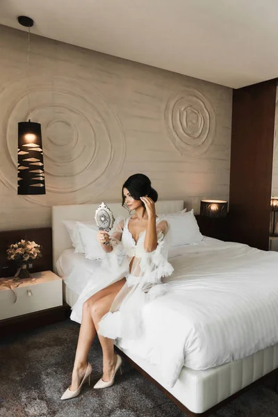 Sexy y hermosa modelo morena chica con un cuerpo perfecto en lencería blanca y peignoir se sienta en la cama, se mira en un espejo y posando en el interior de lujo — Foto de Stock