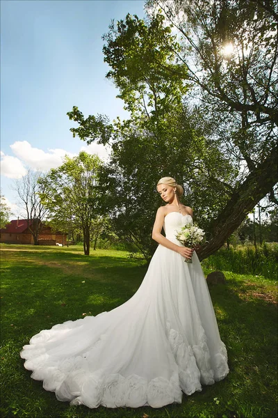 Young fashionable bride, beautiful blonde model girl with stylish wedding hairstyle, in white lace dress with bouquet of flowers in her hands posing outdoors at the sunset