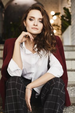 Merdivenlerden ve iç poz güzel ve şık esmer modeli kız mükemmel makyaj, bir şık kırmızı ceket ve trendy damalı pantolon ile oturur