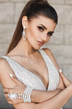 Parlak makyaj ve gülümseyen ve açık havada tuğla duvarın önünde poz parlak gümüş elbisenin içinde düğün saç modeli ile şık ve güzel esmer modeli kız
