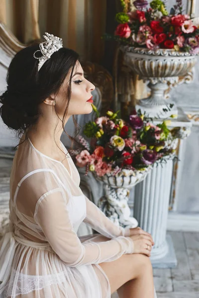 Vackra och moderiktiga brunett modell flicka med ljus makeup och diadem på hennes huvud, i eleganta underkläder och en peignoir sitter på sängen och poserar i lyx vintage inredning — Stockfoto