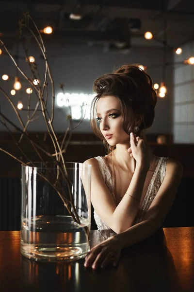 Sexy, busty and fashionable brunette model girl with bright makeup, in stylish dress sits at the table and posing at interior — Stock Photo, Image