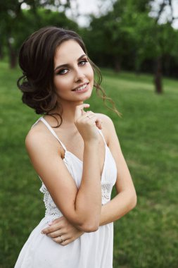 Güzel ve seksi esmer modeli kız parlak makyaj ve şık saç modeli, kısacası beyaz dantel elbise gülümsüyor ve açık havada yeşil park adlı poz