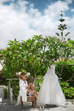 Sarışın güzel, busty ve moda, bir sandalye ve açık havada poz güneşin tadını modeli kız seksi vücut beyaz sabahlığı ve kadın iç çamaşırı, şık güneş gözlüğü ile oturur