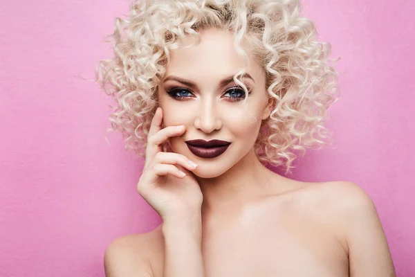 Hermosa y de moda modelo chica con increíbles ojos azules, con el pelo rubio rizado y con maquillaje brillante profesional, sonrisas y posando en el fondo rosa en el estudio — Foto de Stock