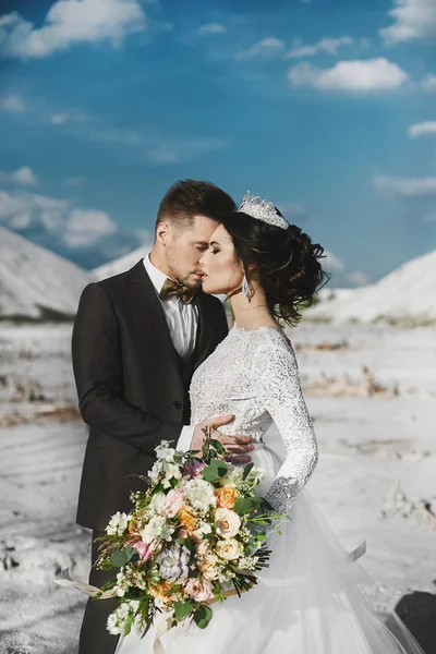 Sexy e bella ragazza modello bruna con acconciatura elegante e con diadema, in abito di pizzo bianco ed eleganti uomini belli in abito alla moda in posa insieme all'aperto al deserto sali — Foto Stock