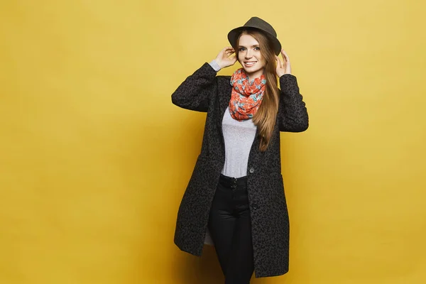 Fashionable and beautiful blonde model girl in stylish coat with leopard patterns and in trendy colorful scarf adjusting her fashionable hat smiling and posing at yellow background — Stock Photo, Image