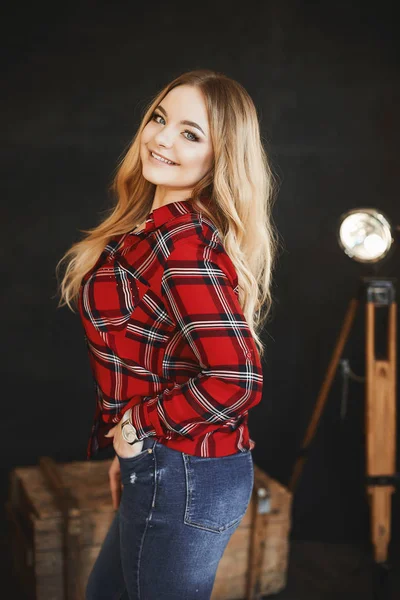 Menina modelo bonita e elegante loira pluss-tamanho com peito grande em camisa xadrez vermelho e em um jeans, fica perto do peito de madeira, sorrindo e posando em estúdio no interior escuro — Fotografia de Stock