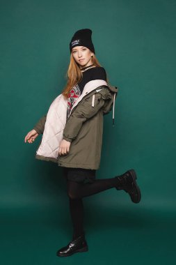 Şık ve güzel genç modeli kız içinde şık aşağı ceket, şık siyah elbise ile renkli yazdırma ve stüdyoda yeşil arka plan, bir siyah şapka poz içinde