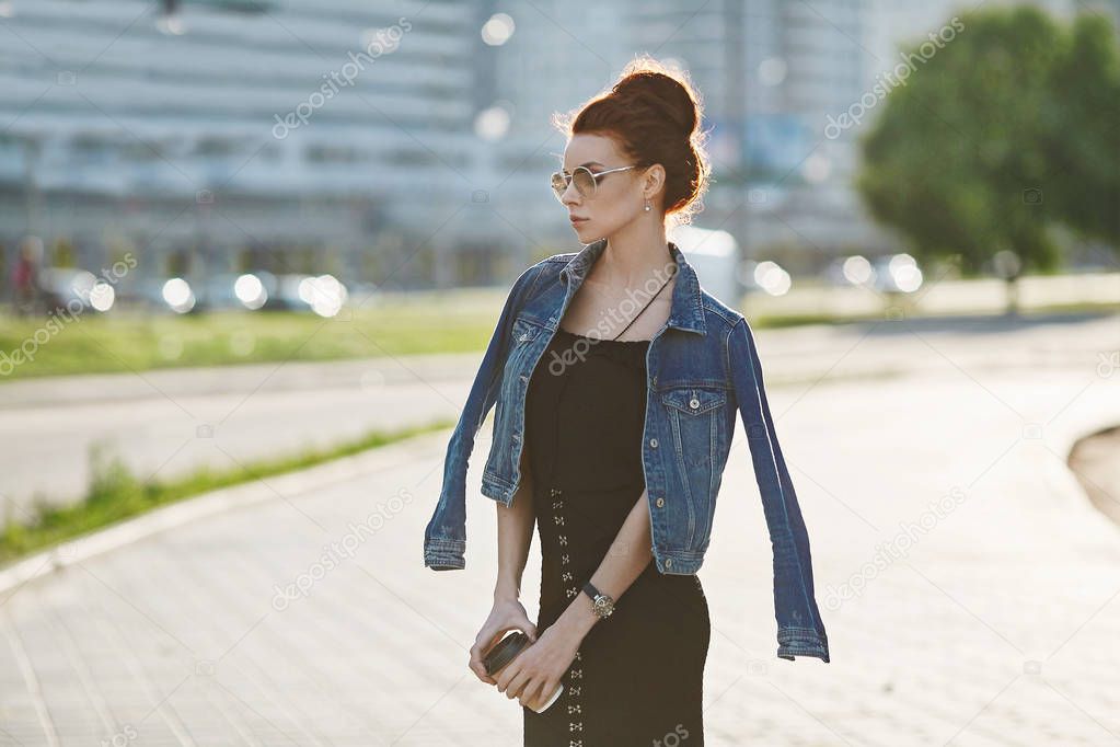 Stylish and beautiful brunette model girl in a jeans jacket, in fashionable black dress and in a trendy glasses, with cup of coffee in her hands posing outdoors at urban background