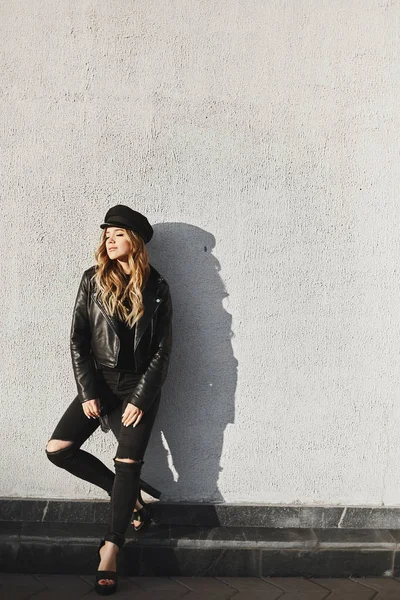 Menina modelo loira elegante e sensual em uma jaqueta de couro preto, em jeans da moda preta e em chapéu elegante sorrindo e posando com olhos fechados ao ar livre — Fotografia de Stock
