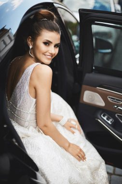 Şık ve seksi esmer modeli kız çıplak sırt, trendy gümüş elbisenin içinde parlak makyaj ile lüks arabada oturuyor gülümseyen ve poz