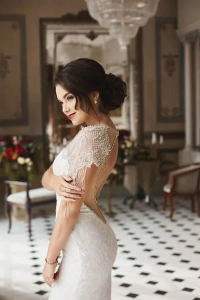 Ragazza sexy e sensuale modello bruna con trucco luminoso e acconciatura alla moda, in un abito elegante in pizzo con schiena nuda, sorridente e in posa a interni vintage di lusso — Foto Stock