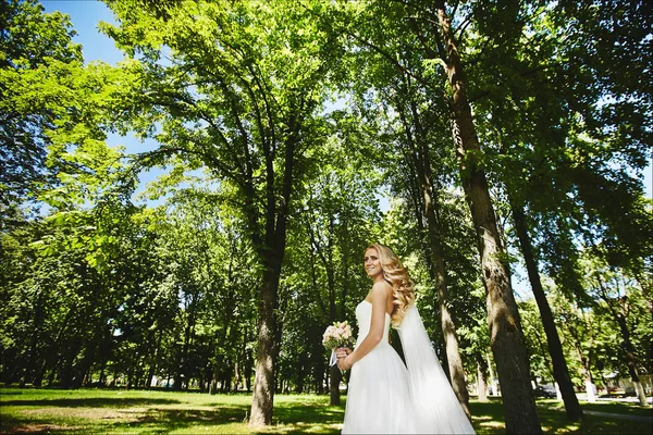 長い白いドレスを着て、結婚式のヘアスタイルと美しい金髪モデルの女の子は公園を散歩して、花の花束を手にポーズ — ストック写真