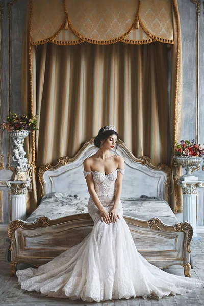 Gyönyörű fiatal menyasszony, szexi barna lány a stílusos és divatos esküvői ruha a meztelen váll ül az ágyon és luxus vintage belső pózol — Stock Fotó