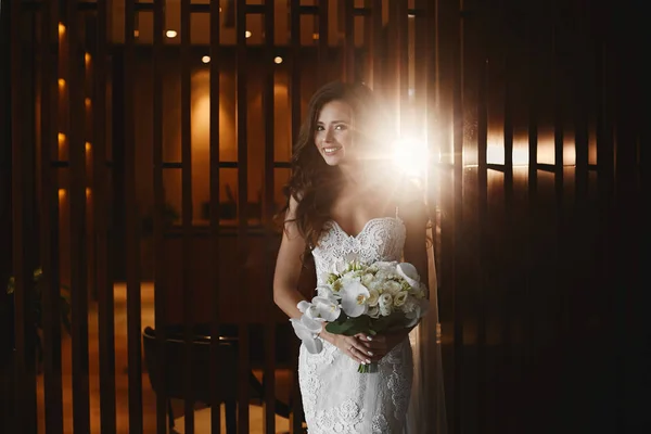 Menina modelo morena bonita, sensual e sexy com maquiagem brilhante em vestido de renda branca elegante com buquê de flores em suas mãos sorrindo e posando em interior elegante de luxo com luz de fundo — Fotografia de Stock