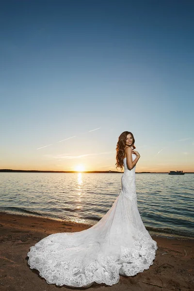 Noiva morena jovem, menina modelo bonita, em vestido de renda branca, está de pé na costa ao pôr do sol — Fotografia de Stock