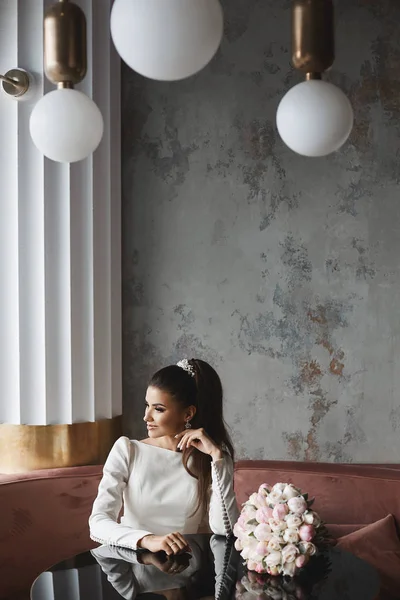 Ragazza alla moda e bella modello bruna con trucco luminoso e con acconciatura da sposa, in abito beige alla moda, si siede a tavola in un caffè, sorridente e in posa all'interno moderno — Foto Stock