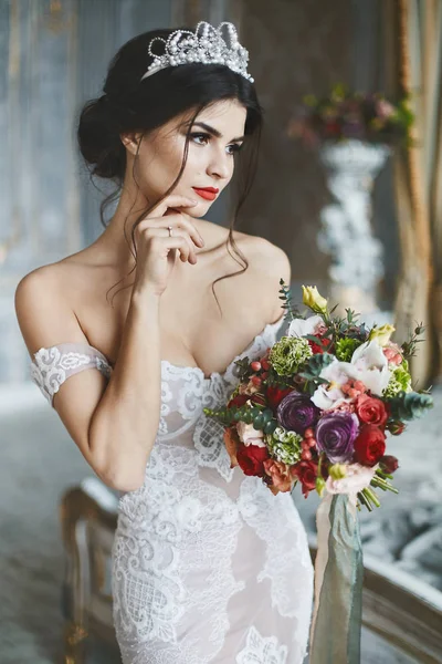 Menina modelo morena sexy e bonita no vestido de noiva elegante e elegante com ombros nus fica com o buquê de flores e posando no interior vintage de luxo — Fotografia de Stock