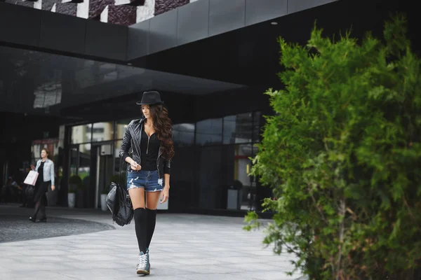 Hermosa, sexy y de moda chica modelo morena con maquillaje brillante, en los pantalones cortos vaqueros, en una chaqueta de cuero y en un sombrero, caminando al aire libre con una elegante mochila negra en la mano — Foto de Stock