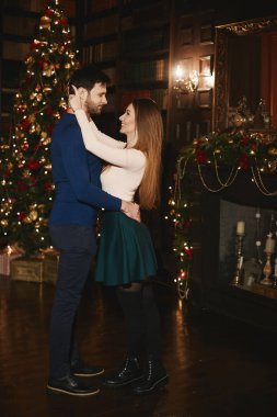 Şık ve güzel bir çift, seksi esmer modeli kız etek ve şık yakışıklı sakallı erkekler hugs ve poz parlak makyaj ile yılbaşı ağacı, yılbaşı için dekore edilmiş iç yakınındaki
