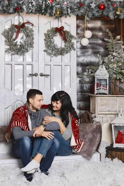 Couple à la mode, beaux hommes élégants étreint la belle fille modèle brune en sweat-shirts gris à la mode, assis et posant près du sapin de Noël à l'intérieur décoré pour le Nouvel An — Photo
