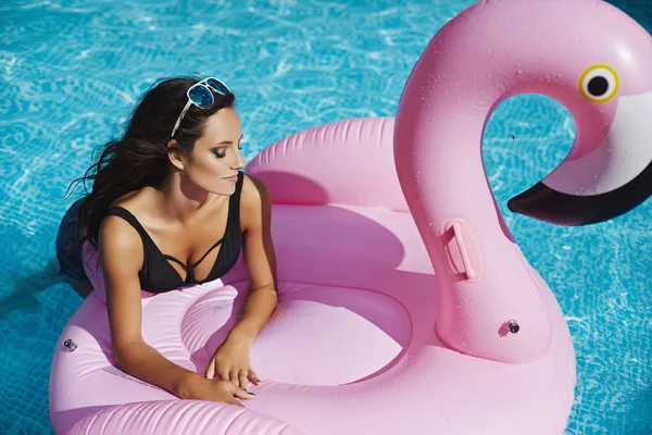 Modische und schöne brünette Model-Mädchen mit perfektem sexy Körper in stylischem schwarzen Bikini und glamouröser Sonnenbrille, posiert auf einem aufblasbaren rosa Flamingo am Pool im Freien — Stockfoto