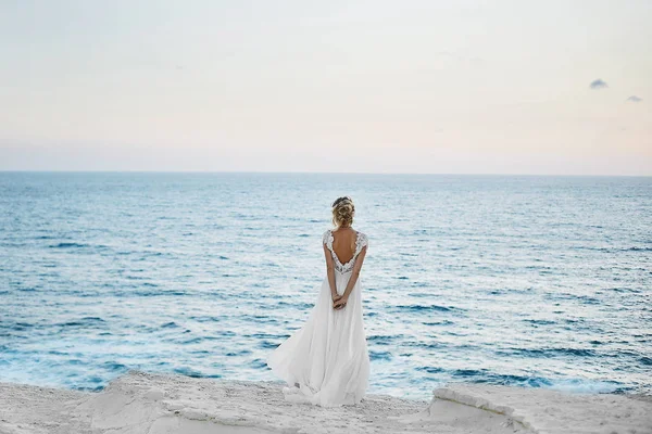 Bella giovane ragazza modello bionda in abito bianco si erge indietro e guarda il mare — Foto Stock