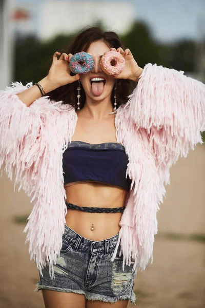 Fröhliche und modische Brünette Modell Mädchen mit perfektem sexy Körper in Jeans Shorts und in einem Top, Spaß haben und posiert mit einem leckeren Donuts in ihren Händen — Stockfoto